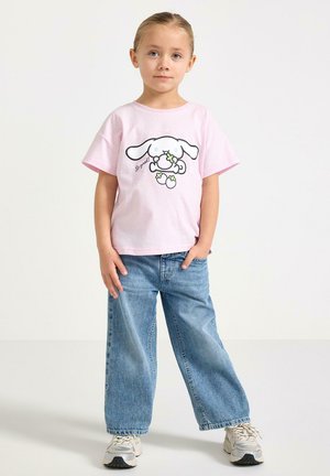 Ung pige står med hånden i lommen, iført en lyserød T-shirt med hundemotiv, blå vidbenede jeans og hvide sneakers.