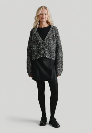 Blondynka w czarnej sukience, czarnych rajstopach, czarnych butach i czarno-białym swetrze o grubym splocie stoi na tle jednolitego tła.