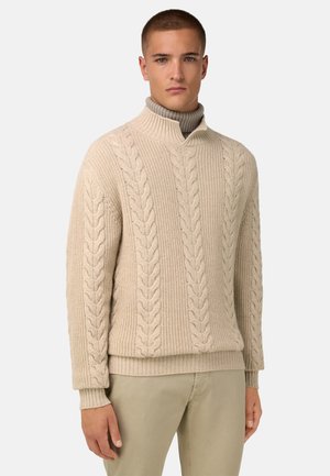 Beiger Kabelstrickpullover mit hohem Kragen und gerippten Bündchen. Grobe Textur und vertikales Muster, kombiniert mit hellen Hosen.