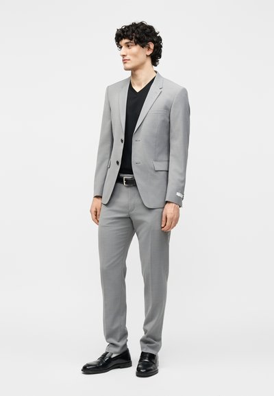 Νέος άνδρας με σγουρά μαλλιά φοράει ανοιχτογκρι κοστούμι, μαύρο μπλουζάκι με V-λαιμόκοψη, μαύρη ζώνη και μαύρα παπούτσια και στέκεται μπροστά σε λευκό φόντο.