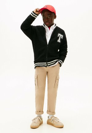 Ragazzo con berretto rosso, giacca nera con zip e lettera T, maglietta bianca, pantaloni cargo beige e sneakers beige, in piedi con una mano in tasca e l'altra che si aggiusta il berretto.