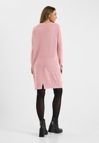Roze gebreide jurk met lange mouwen, zijslitsen en een relaxte pasvorm. In combinatie met zwarte enkellaarsjes met dikke hakken.