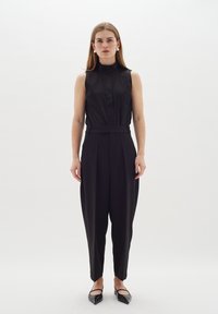 Blusa senza maniche nera con scollatura a bottoni, abbinata a pantaloni neri a vita alta e svasati; il modello indossa ballerine appuntite.