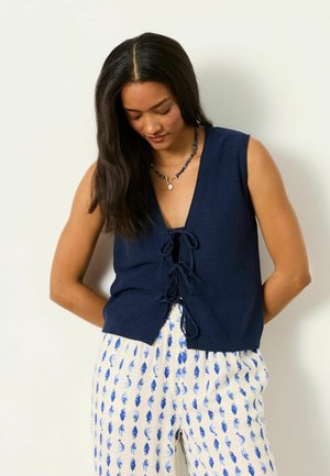 Woman with long dark hair wearing a navy sleeveless knit vest tied in front and white pants with blue leaf prints, looking down.