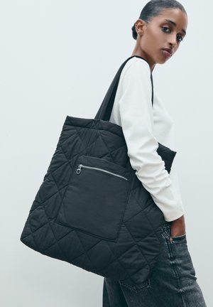 Schwarze gesteppte Tragetasche mit einer Reißverschlusstasche auf der Vorderseite, die ein Diamantmuster aufweist, robuste Griffe hat und aus einem glatten Stoff gefertigt ist.