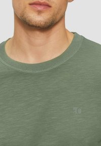 Zielony sweter typu crewneck wykonany z lekkiego materiału, z delikatną teksturą i małym logo nadrukowanym na klatce piersiowej.