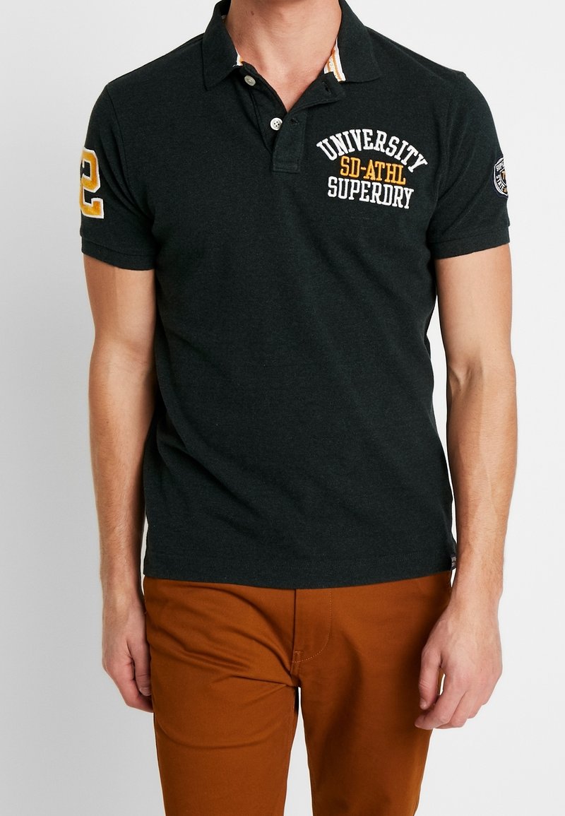 Mørkegrå polo-shirt med klassisk krave, broderet "Universitet" og nummer 2 på ærmet, parret med brune bukser.