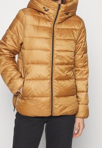 Kvinde iført en tan farvet quilted puffer jakke med en frontlynlås og sidelommer, parret med sorte bukser.