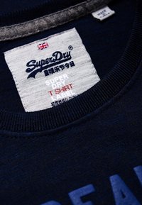 Etiqueta marki SuperDry z flagą Wielkiej Brytanii na granatowym T-shircie, z japońskim napisem i tekstem "SUPER DRY T SHIRT LABEL".