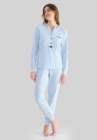 Mujer de pie descalza, vistiendo una blusa de manga larga azul claro y pantalones con pequeños motivos florales en azul oscuro, sobre un fondo liso.