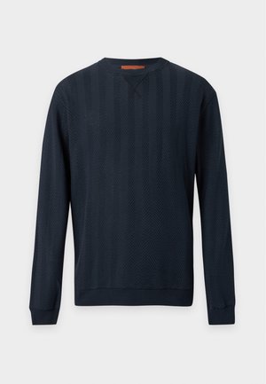Homme portant un pull noir texturé, un pantalon gris, des chaussures en cuir noires et des lunettes de soleil foncées, debout les mains dans les poches.