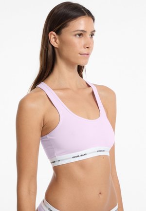 Soutien-gorge de sport rose clair avec des bretelles larges, un col arrondi et un bandeau élastique blanc comportant le nom de la marque en noir. Matière lisse et extensible.