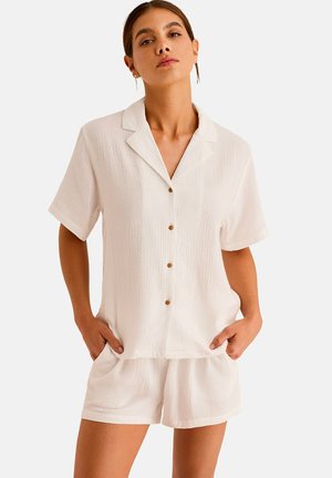 Femme portant une chemise blanche à manches courtes texturée et un short assorti, debout les mains dans les poches devant un fond uni.