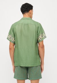 Camisa verde de manga corta con patrones de espirales bordados en las mangas, tejido ligero con un acabado texturizado y un diseño de cuello.