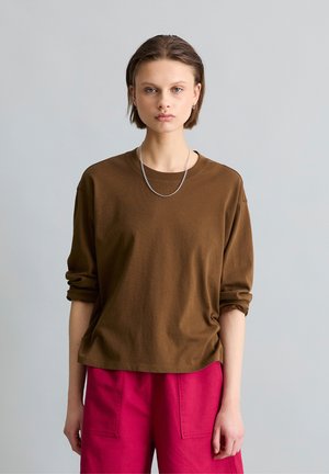 Młoda osoba z krótkimi włosami nosi brązową koszulę z długim rękawem, srebrny naszyjnik w formie łańcuszka oraz jaskrawo różowe spodnie, stojąc na jednolitym jasnym tle.