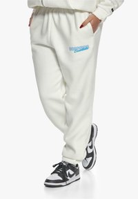 Kremfargede joggebukser med elastisk midje, sidelommer og en blå logo med teksten "dropsize essentials." Kombinert med svarte og hvite joggesko.