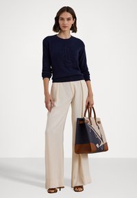 Ναυτικό πλεκτό πουλόβερ με ανάγλυφο λογότυπο, συνδυασμένο με κρέμα παντελόνια σε φαρδιά γραμμή και μια τσάντα tote σε δύο τόνους με ναυτικό φόντο και καφέ λεπτομέρειες.