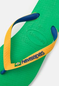 Green rubber flip-flop with a textured sole, featuring yellow and blue straps. "Havaianas" logo printed on strap with a small Brazilian flag accent.