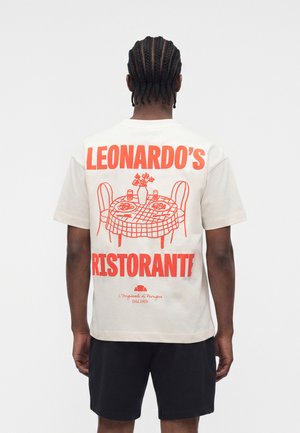 Muž s vrkočmi, ktorý má na sebe biele tričko s červenou potlačou jedálenského stola a nápisom „Leonardo's Ristorante“ na chrbte.