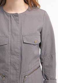 Mujer vistiendo una chaqueta gris con bolsillos tipo solapa y cremalleras, mostrada de los hombros a la cintura sobre un fondo blanco.