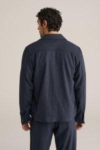 Tmavomodrý vlnený kabát s voľným strihom, vybavený špicatým golierom, gombíkovými manžetami a hladkou textúrou. Pohľad zozadu zobrazuje švové detaily.