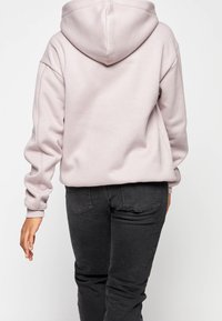 Hellrosa, übergroßer Kapuzenpullover mit einer geräumigen Kapuze, gerippten Bündchen und geradem Saum. Kombiniert mit dunkelgrauen, engen Jeans.