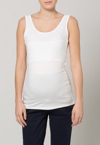 Nais nainen, joka pukeutuu valkoiseen hihattomaan äitiyspaitaan ja tummiin housuihin, seisoo yksinkertaista harmaata taustaa vasten.