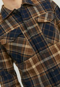 Camisa de franela a cuadros en marrón y azul marino con dos bolsillos en el pecho, botones y una tela texturizada con líneas en colores más claros que se intersectan.