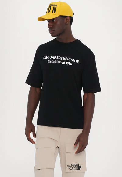 Schwarzes Kurzarm-T-Shirt mit weißem Text "DSQUARED2 HERITAGE Established 1995." Kombiniert mit beigen Cargo-Hosen mit Taschen.