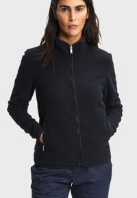 Femme aux longs cheveux foncés portant une veste polaire noire zippée et un pantalon bleu marine, mains dans les poches de la veste, expression neutre.