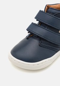 Marineblaue Lederschuhe mit zwei verstellbaren Klettverschlüssen, weißer Gummisohle, glatter Oberfläche und kontrastierendem, tanfarbigem Innenfutter.