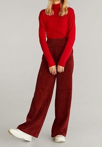 Un pull à col roulé rouge associé à un pantalon en velours côtelé bordeaux à jambes larges. Le modèle porte des baskets blanches, affichant une coupe décontractée et une texture.