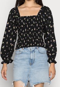 Blouse noire à motifs floraux avec des manches bouffantes et décolleté carré, présentant un design smocké. Assortie à une jupe en denim bleu clair.