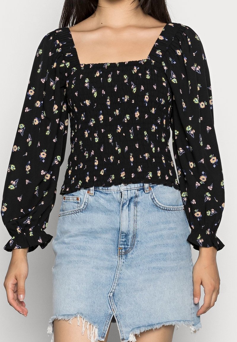 Blouse noire à motifs floraux avec des manches bouffantes et décolleté carré, présentant un design smocké. Assortie à une jupe en denim bleu clair.