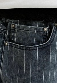 Gros plan sur un jean en denim foncé avec des fines rayures blanches verticales et des rivets métalliques sur les bords des poches.