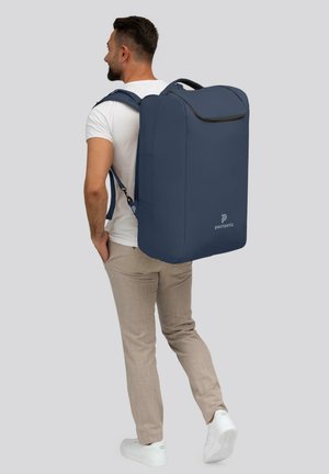 Uomo con pantaloni beige e sneakers bianche che porta uno zaino rettangolare grande di colore blu navy etichettato "pactastic" su sfondo grigio.