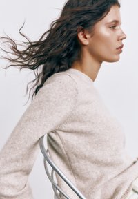 Mladá žena s dlhými vlnitými vlasmi, oblečená v béžovom svetri, sedí bokom na kovovej stoličke pred jednoduchým pozadím.