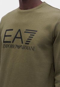 Homme portant une chemise à manches longues vert olive avec un grand logo noir EA7 Emporio Armani imprimé sur la poitrine.
