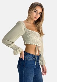 Hellbeiges, verkürztes Top mit Raffung, langen Ärmeln und einer Bindeschleife an der Vorderseite. Kombiniert mit dunkelblauen, hochgeschnittenen Jeans.