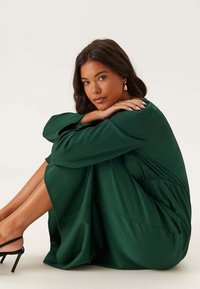 Abito verde fluido con maniche lunghe, morbida consistenza e vestibilità ampia. Modella seduta con le braccia avvolte intorno alle gambe, indossando semplici tacchi neri.