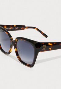 Skildpaddemønstrede firkantede solbriller med gradient grå linser og gyldent "TH" logo på brillestangen, vippet på hvid overflade.