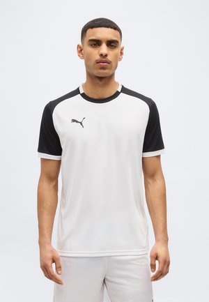 Moški, ki nosi belo in črno športno majico Puma ter bele kratke hlače, stoji obrnjen proti naprej z nevtralnim izrazom na obrazu.