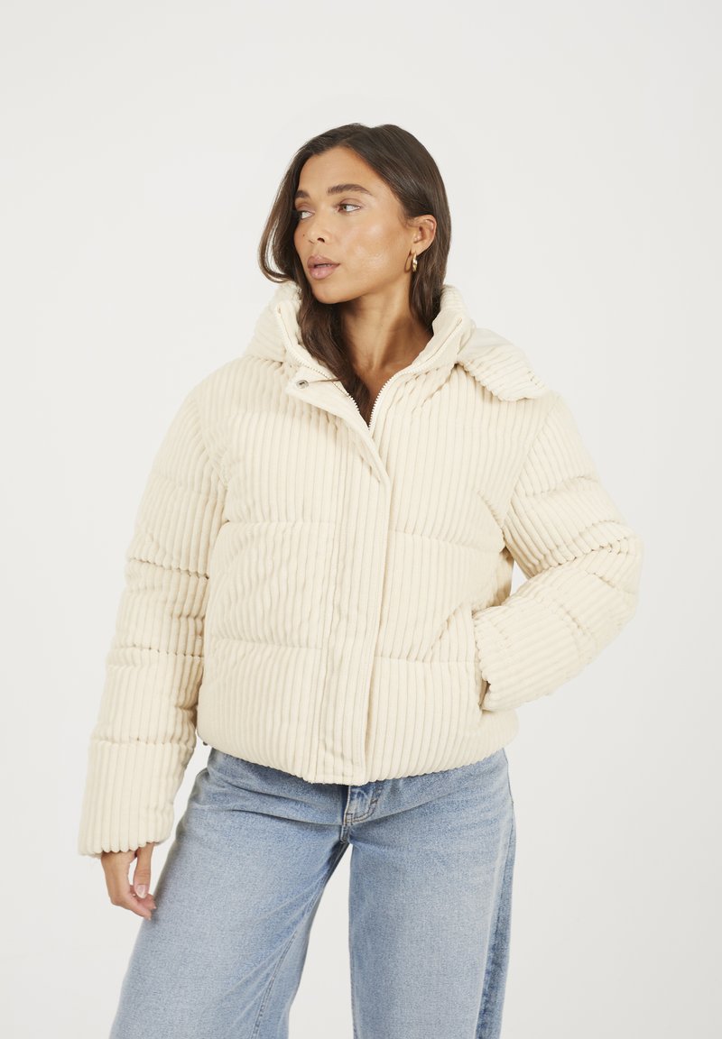 Veste en duvet en tissu côtelé crème avec un col montant, fermeture éclair à l'avant et deux poches latérales. Portée sur un pantalon en denim bleu clair.