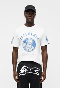 Λευκό μπλουζάκι με μπλε γραφικό τύπωμα μιας γυναίκας και το κείμενο "ICECREAM" από πάνω. Μαύρο παντελόνι με λευκό καλλιτεχνικό σχέδιο σκύλου στο πίσω μέρος.