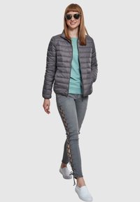 Jachetă căptușită gri, bluză teal, blugi gri cu detalii laterale din șnur, și pantofi albi slip-on. Model purtând ochelari de soare reflectanți, în picioare.