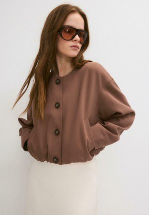 Femme aux longs cheveux bruns portant de grandes lunettes de soleil marron et une veste marron boutonnée, les mains dans les poches, devant un mur uni.