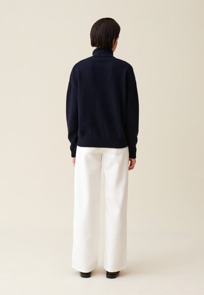 Pulover turtleneck bleumarin cu o croială lejeră și tiv ribflat, asortat cu pantaloni albi cu croială largă și pantofi negri. Fond simplu.