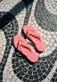 Tongs rose vif posées sur un sol en mosaïque noir et blanc avec des motifs incurvés et une ombre projetée sur une partie de la scène.