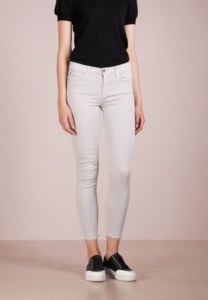 Schmal geschnittene weiße Jeans mit ausgefransten Säumen, getragen zu einem schwarzen Kurzarmoberteil und schwarz-weißen Plateau-Sneakern, stehend auf beigem Boden.