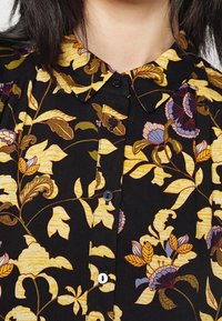Camisa negra con un patrón floral en amarillo, morado y verde, abotonada hasta el cuello, llevada por una persona con cabello oscuro.